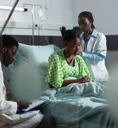 African american medical team working on healing patient in hospital ward bed. Man and woman with doctors occupation examining young adult for treatment using monitor and stethoscope